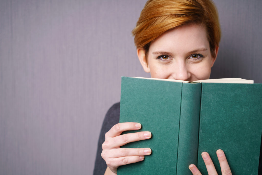 Eine Frau hält ein Buch vor ihr Gesicht und lächelt. (freiwillige Quarantäne)
