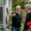 Energiesparen: 2 Männer stehen draußen im Garten vor einem Haus und schauen die installierte Klimaanlage an.